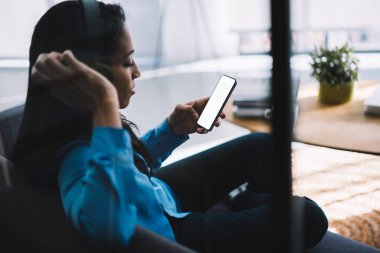 Evde boş ekranla akıllı telefon sörfü yaparken gündelik kıyafetleriyle kanepeye oturmuş kulaklık takan genç etnik modern kadının yan görüntüsü.