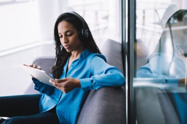 Uzun siyah saçlı, odaklanmış, kulaklıkla müzik dinleyen ve koltukta otururken büyük tablet üzerinde çalışan etnik bir kadın.