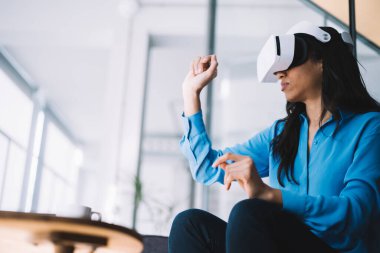 Mavi gömlekli kadın uzmanın yan görüntüsü VR aygıtını test ederken el ve yumruk kaldırılmış bir şekilde masada oturuyor.