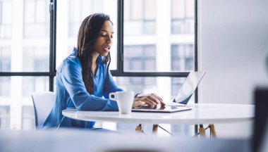 Mavi bluzlu, yansıtıcı Afrikalı Amerikalı kadının seçici odak noktası kahveden zevk alması ve modern ofisteki yuvarlak masada dizüstü bilgisayar kullanması.