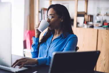 Düşünceli, uzun saçlı, Afro-Amerikalı bir kadın laptopla çalışırken ara verip kahve içiyor.