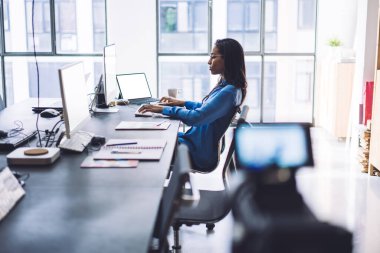 Modern iş yerinde boş ekranları olan cihazlar üzerinde çalışırken, ciddi etnik kadın yöneticilerin ofis hayatını tripod kamerayla kaydetmesinin yan görüntüsü