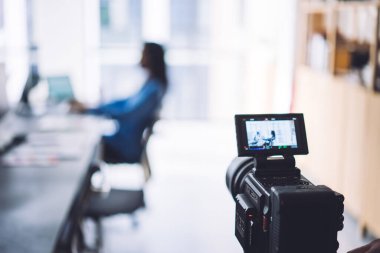 Dijital ekranlı modern video kamera, yumuşak odaklı arka planda bilgisayar masasında oturan tanınmayan işçiyi gösteriyor.
