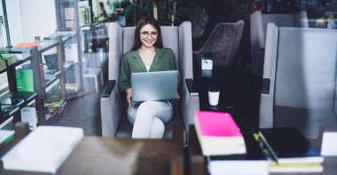 Modern kütüphane odasındaki rahat koltukta gülümseyen ve dizüstü bilgisayar kullanan çekici genç bayan kameraya bakıyor.