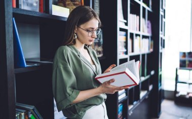 Öğrenci kitap raflarına yaslanarak özenle kitap okuyor.