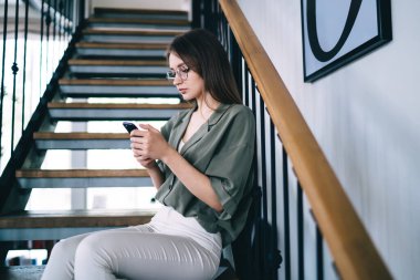 Moda kıyafetleri ve modern yuvarlak bardaklarla dekoratif merdivenlerde dinlenen ve içi aydınlık olan bir odada akıllı telefon kullanan kadının yan görüntüsü.
