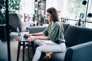 Konsantre olmuş düşünceli bir kadının, rahat koltukta oturmuş, elinde bir bardak içecekle modern odada dizüstü bilgisayar kullanışının yan görüntüsü. 