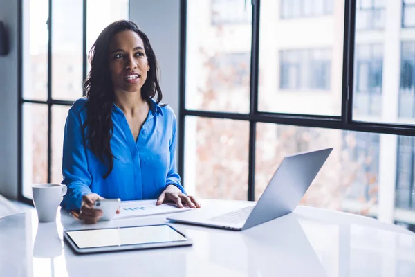 Reflective African American woman in blouse messaging smartphone and browsing laptop and tablet while sitting at desk in office on blurred background
