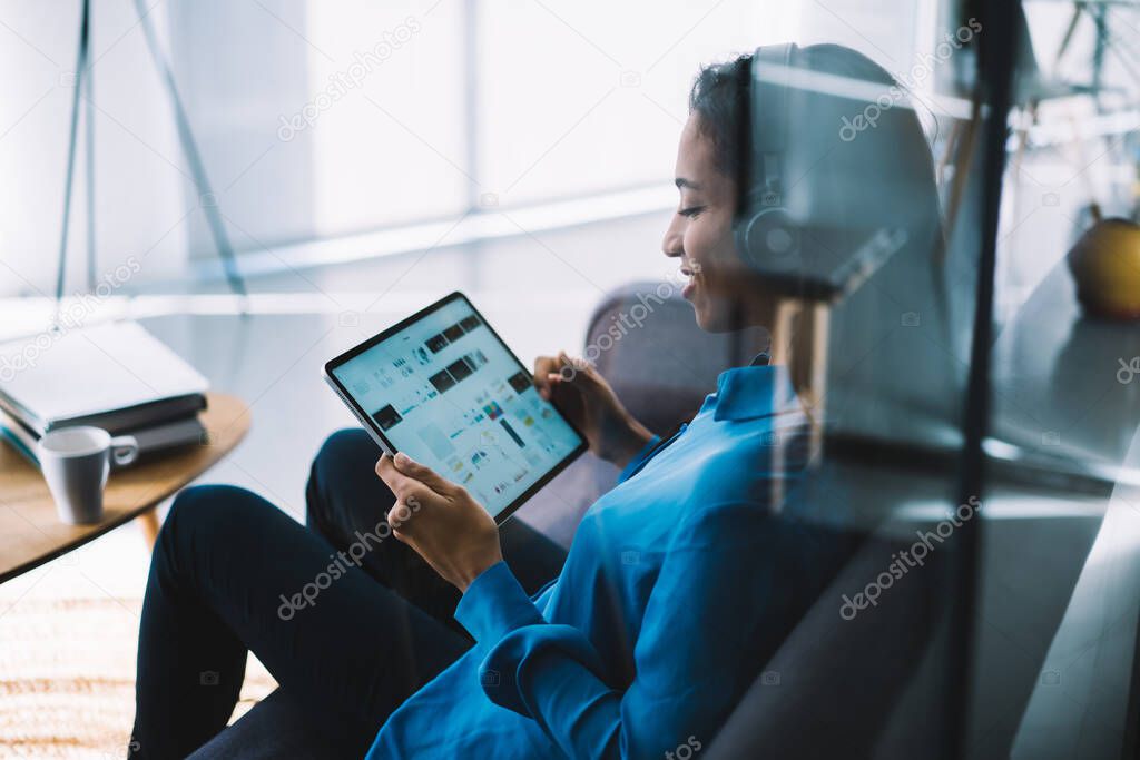 Side view through glass of casual cheerful woman in headphones surfing tablet with empty screen while sitting on sofa beside coffee table