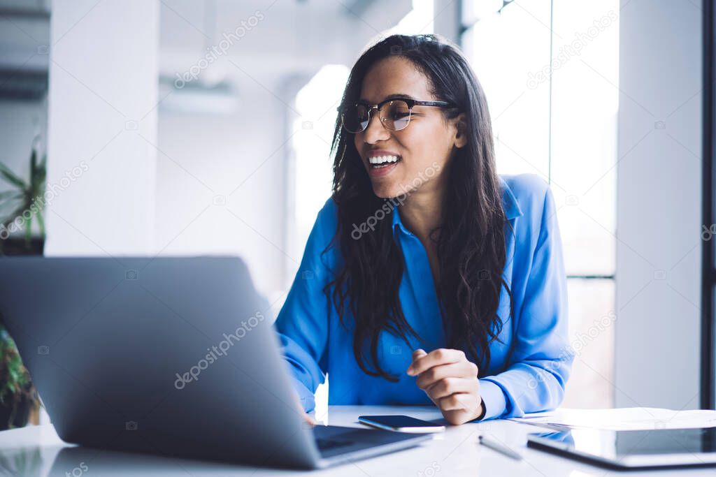 Cheerful African American businesswoman in glasses browsing information on laptop reading good news and sitting at round table in light cafe