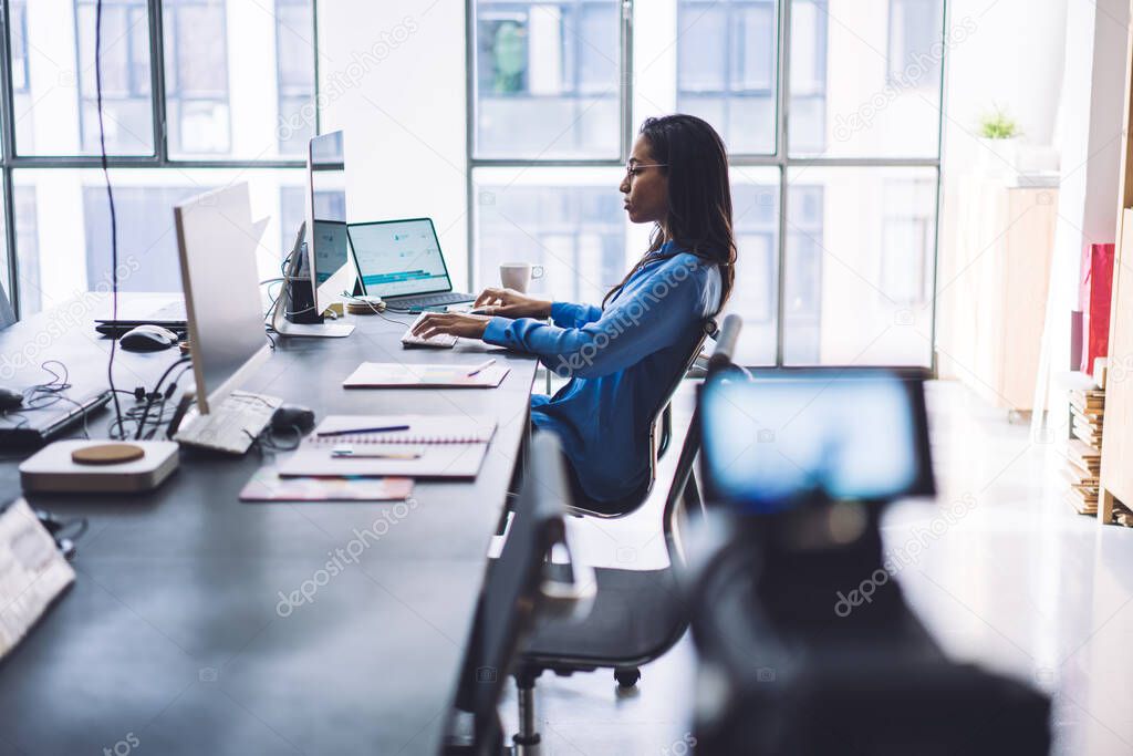 Side view of confident black businesswoman shooting on video camera work process while browsing gadgets at table in modern office