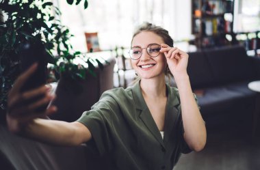 Çekici, zeki, gözlük takan ve cep telefonuyla selfie çeken, boş kütüphanede gülümseyen bir öğrenci.