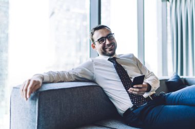 Kafkasyalı başarılı patronun portresi modern dijital telefonla koltukta dinleniyor. Olumlu, olgun bir adam resmi olarak kameraya gülümsüyor ve akıllı telefon cihazıyla iş görüşmelerini bekliyor.