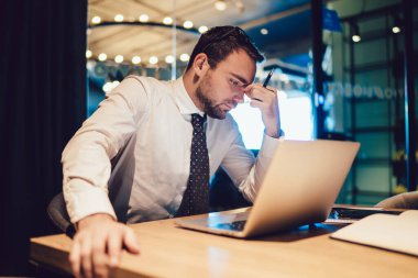 Gündelik yorgun ofis çalışanı 4G kablosuz bağlantılı dizüstü bilgisayar kullanarak masa başında oturuyor ve fazla mesai yapıyor, Kafkasyalı işadamı teslim tarihi boyunca sorunları çözmek için resmi olarak giyinmiş.