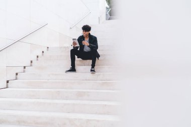 Gündüz vakti şehir merdivenlerinde otururken günlük kıyafetleriyle gezen şık, neşeli, genç, Afro-Amerikan bir adam.