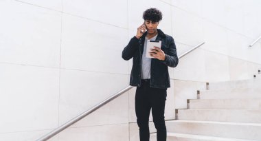 Modern tarz, ciddi, esmer, Afro-Amerikan bir adam. Günlük kıyafetleriyle telefonda konuşuyor ve tablet kullanıyor. Gün ışığında şehir merdivenlerinde dikilirken.