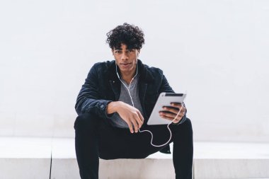 Merdivenlerde oturmuş kulaklıkla müzik dinleyen ve beyaz duvara bakan Afro-Amerikalı genç yakışıklı adam.