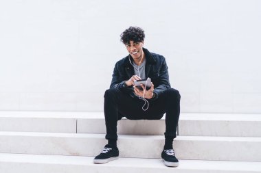 Tabletle gülümseyen ve merdivenlerde oturmuş kulaklıkla müzik dinleyen ve beyaz duvara bakan Afro-Amerikalı genç yakışıklı adam.