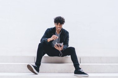 Beyaz merdivenlerde gülümseyen, kulaklıkla müzik dinleyen ve beyaz duvara bakan Afro-Amerikalı genç bir erkek.