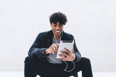 Neşeli Latin hippi adam elinde dokunmatik bir kağıt tutarken ve sosyal ağlara paylaşım için içerik metni yazarken aynı zamanda internet sitesine göz gezdirirken elektronik kulaklıklı adam ses şarkılarını seçiyor.