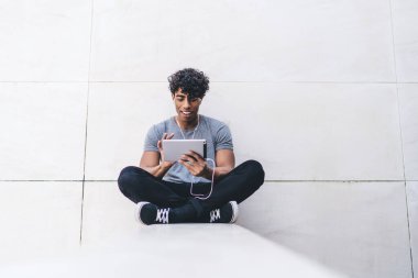 Gülümseyen, yakışıklı, sıradan giyinmiş, bacak bacak bacak üstüne atmış, dışarıda kulaklık takmış, tablette film izleyen siyah bir erkek.