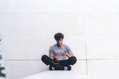 Gülümseyen, yakışıklı, sıradan giyinmiş, bacak bacak bacak üstüne atmış, dışarıda kulaklıklarla fotoğraf çeken siyah bir erkek.