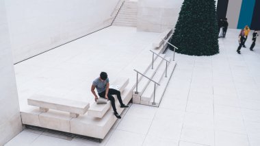 Modern etnik adam, elinde kağıtlarla bankta otururken ve iş merkezinin çağdaş beyaz lobisinde cihaz kullanırken dizüstü bilgisayarına göz atıyor.