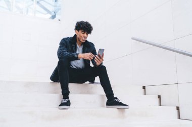 Beyaz modern binada merdivenlerde otururken kıvırcık saçlı, mutlu, Afro-Amerikalı bir adam akıllı telefon kullanıyor.