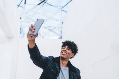 Kıvırcık saçlı, modaya uygun kot elbiseli, akıllı telefonlu, parlak bir odada selfie çeken ve gülümseyen Amerikalı 'ya sevindim.