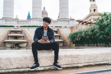 Genç Afro-Amerikalı gözlüklü ve elinde tabletle parktaki çeşmenin kenarında oturan siyah elbiseli bir adam.