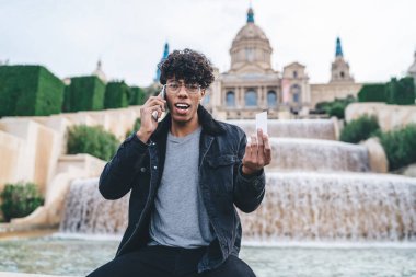 Hayretler içinde genç Afro-Amerikan bir erkek, günlük giysiler içinde elinde reklam kartıyla oturuyor ve su tesislerinin kenarında cep telefonuyla konuşuyor.