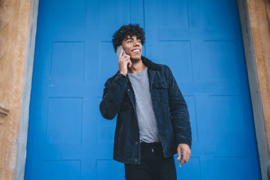 Gülümseyen kıvırcık, siyah ceketli ve kot pantolonlu yetişkin bir erkeğin sokak mavi kapısının önünde dikilip gözlerini kaçırdığı düşük açı. 