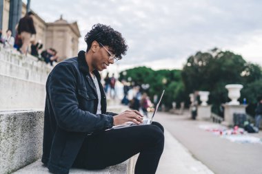 Gözlüklü neşeli etnik erkeğin şehir sokaklarında arka planda insanlarla merdivenlerde otururken keyfi yerinde dizüstü bilgisayara bakması.