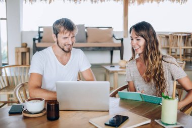 Gülümseyen başarılı genç serbest çalışanlar, rahat, ahşap iç kısımları olan bir kafede oturup not alırken dizüstü bilgisayarlarında iş birliği yapmaktan zevk alıyorlar.