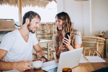 Gülümseyen uzaktan kumandalı genç çalışanlar kafede oturmuş kahve içiyor ve akıllı telefonlarda gösteri yaparak işle ilgili fikirleri paylaşıyorlar. 