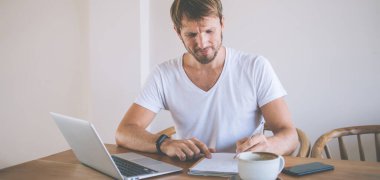 Düşünceli, gülümseyen, günlük, traşsız, not defterine yazan ve masada kahve fincanıyla otururken dizüstü bilgisayar kullanan bir adam.