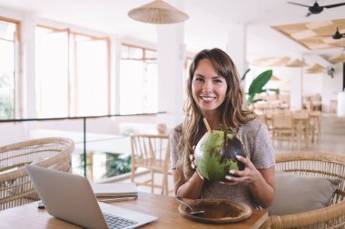 Gündelik giysiler içinde gülümseyen güzel bir kadın dizüstü bilgisayar ve defterle masada dinleniyor ve kafede doğal içeceklerin tadını çıkarıyor ve bulanık arka plandaki kameraya bakıyor.