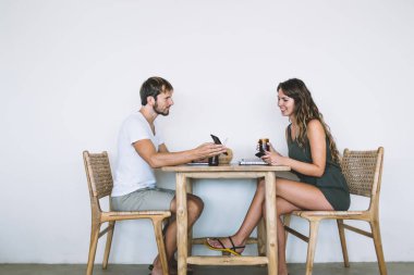 Yetişkin, yakışıklı, sakallı, günlük kıyafetlerle gezen ve kafeteryada beyaz arka planda giyinmiş güzel bir bayana akıllı telefon ekranında fotoğraflar gösteren erkek.