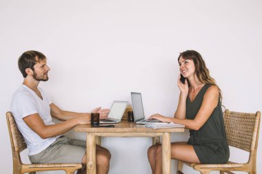 Sıradan giyinmiş, yakışıklı, sakallı bir erkeğin dizüstü bilgisayarda sörf yaparken beyaz arka planda akıllı telefondan konuşan neşeli, güzel bir kadının yan görüntüsü.