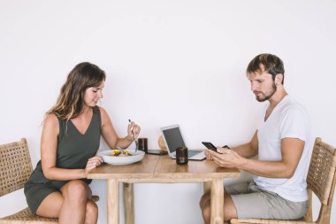 Günlük giysili bir adam telefona dikkatle bakarken, kısa elbiseli bir kadın kahvaltısını yapıyor, ikisi de mutfakta oturuyor ve hafta sonu sabahının tadını çıkarıyor.