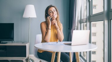 Positif kadın serbest çalışanın portresi evde oturmuş kameraya gülümserken cep telefonu cihazıyla müşteri hizmetlerine sesleniyor. 30 'lu yaşlardaki mutlu hippi kız dedikodu için arkadaşına telefon ediyor.