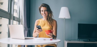 Gülümseyen ve kameraya bakan başarılı kadın serbest çalışan bir fincan çay ve akıllı telefonla dinlenirken yuvarlak masada dizüstü bilgisayarla oturuyor.