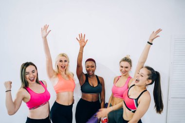 Antrenman stüdyosunda spor malzemeleriyle kazandıkları zaferi kutlarken bir araya gelip kollarını havaya kaldıran bir grup mutlu çok ırklı kadın.