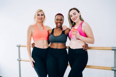 Arkadaş canlısı ve çok ırklı spor kadınlarından oluşan bir şirket beyaz arka planda, bale barında kameraya sarılıp bakarken...