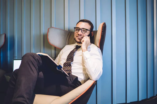 Portrait of man professional business worker dressed in formal wear talking with partner via mobile phone and using textbook while sitting indoors, Caucasian male intelligent lawyer phoning via app