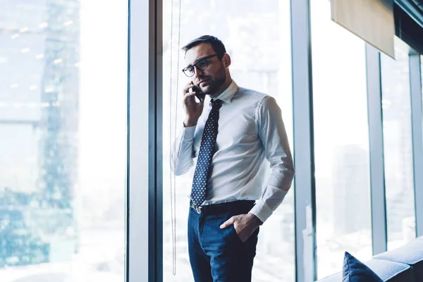 Confident male owner of company calling to business partner via smartphone device for communicate about project, handsome man in optical eyewear making conversation with colleague for consultancy