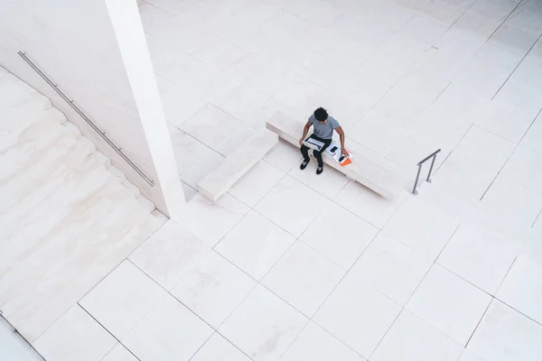 From above contemporary white tiled office hallway with male freelancer in casual clothes using laptop working with papers in daylight