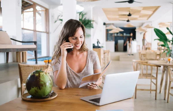 Yaz günü masada hindistan cevizi içeceği ve dizüstü bilgisayarla otururken akıllı telefondan konuşan ve not alan sıradan, rahat, genç bir kadın.