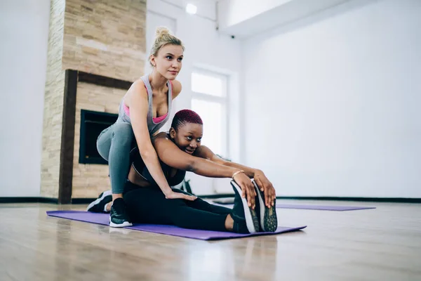 Young blonde female helping cheerful black haired African American friend with stretching, both looking in mirror and enjoying going to yoga classes together