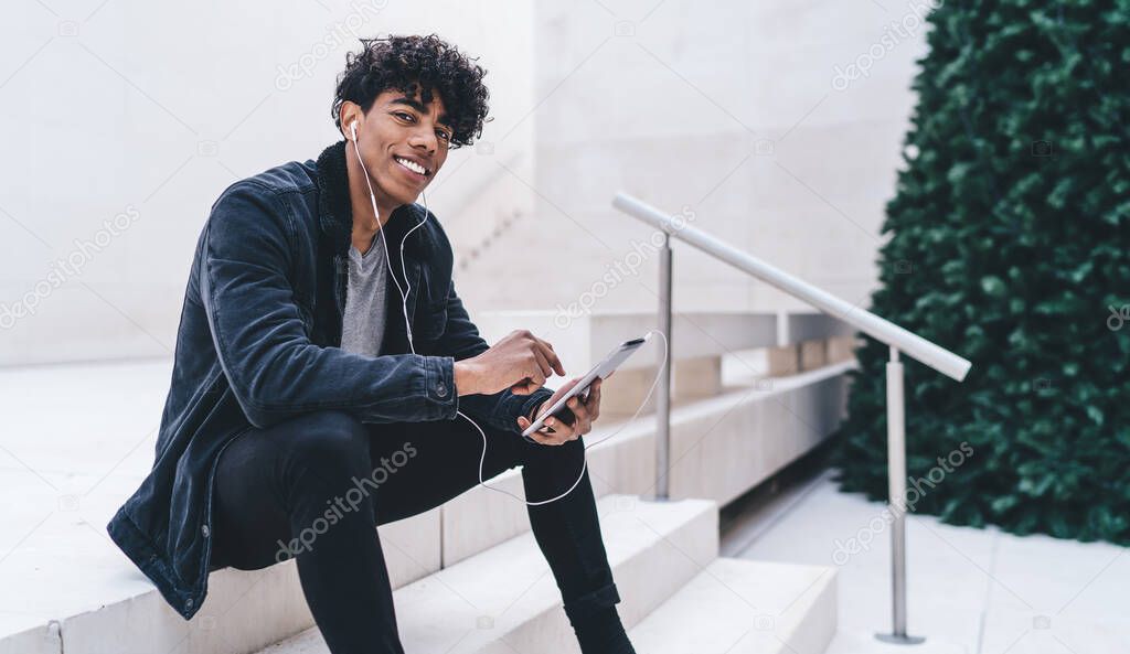 Young arrogant African American male student wearing black clothes sitting on stairs with tablet listening to music smiling and looking at camera